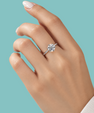 A close-up of a hand wearing a silver-toned diamond ring with a round cluster setting, set against a solid light teal background.