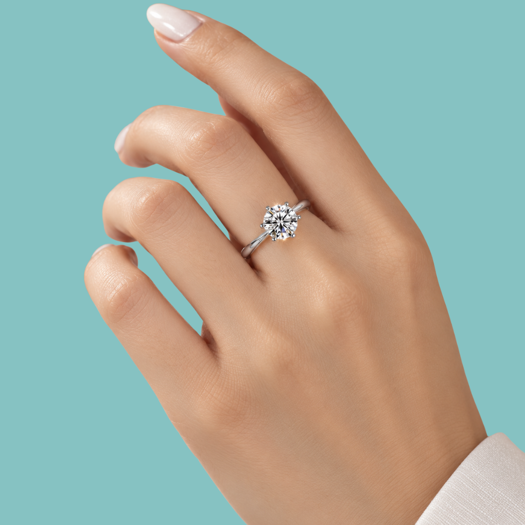 A close-up of a hand wearing a silver-toned diamond ring with a round cluster setting, set against a solid light teal background.