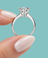 A silver solitaire engagement ring with a round brilliant-cut diamond held between two fingers against a solid teal background.