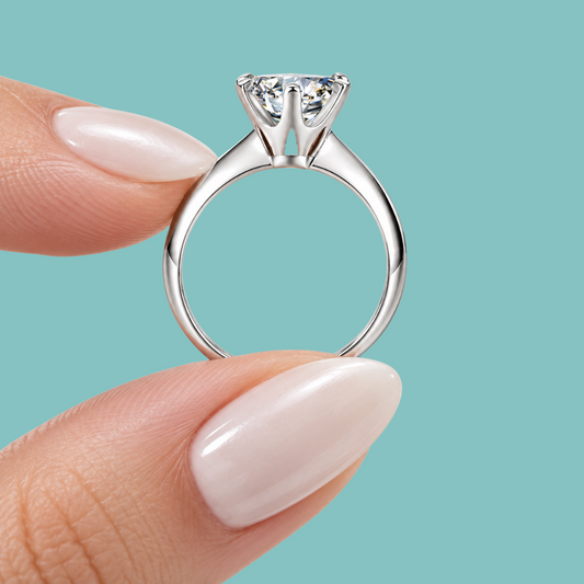A silver solitaire engagement ring with a round brilliant-cut diamond held between two fingers against a solid teal background.