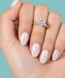 A close-up of a hand with glossy pale pink nails wearing a gold ring with a round clear gemstone, set against a solid teal background.