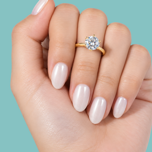 A close-up of a hand with glossy pale pink nails wearing a gold ring with a round clear gemstone, set against a solid teal background.