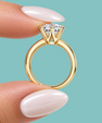 A close-up of a gold ring with a brilliant-cut clear gemstone held between two fingers with pale pink manicured nails, set against a solid light teal background.