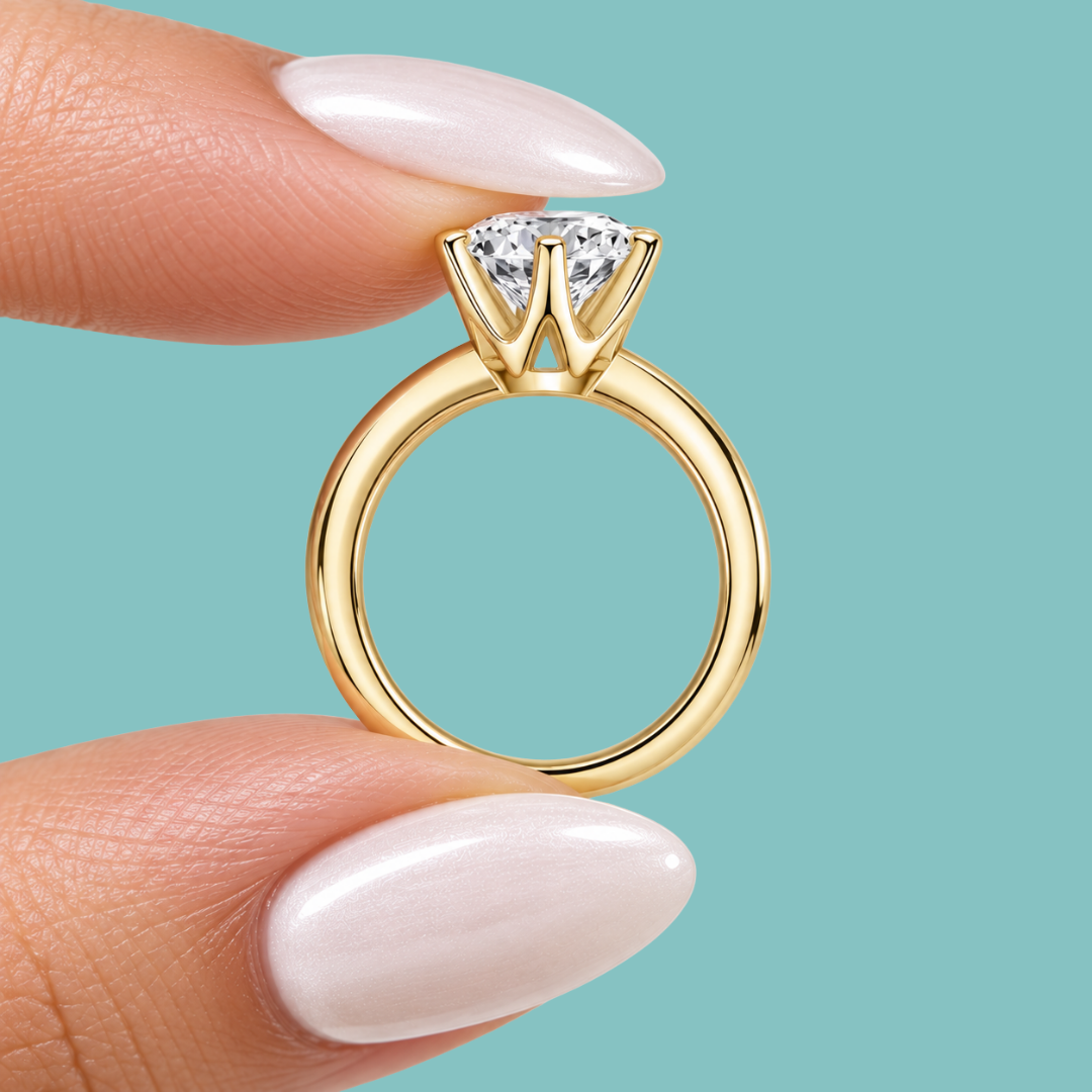A close-up of a gold ring with a brilliant-cut clear gemstone held between two fingers with pale pink manicured nails, set against a solid light teal background.