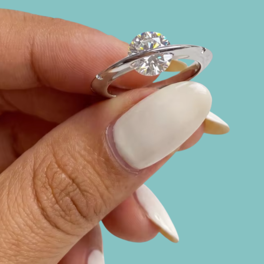 Saturn’s Kiss moissanite ring featuring a round centre stone crossed by a polished silver-toned band, with small side stones.