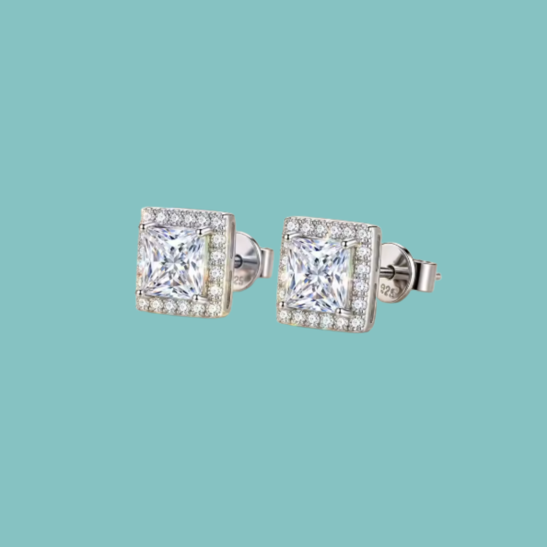 A pair of square stud earrings with princess-cut clear gemstones surrounded by a halo of smaller clear stones, set in silver-toned metal on a solid light teal background.