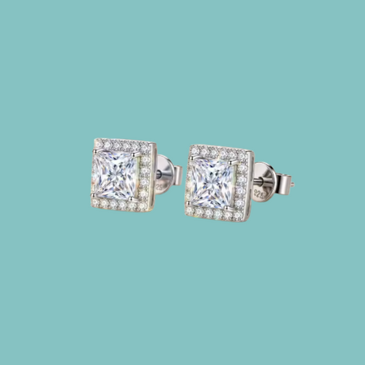 A pair of square stud earrings with princess-cut clear gemstones surrounded by a halo of smaller clear stones, set in silver-toned metal on a solid light teal background.