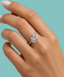 A close-up of a hand wearing a silver ring with a marquise-cut diamond and an emerald-cut diamond, set against a solid teal background.