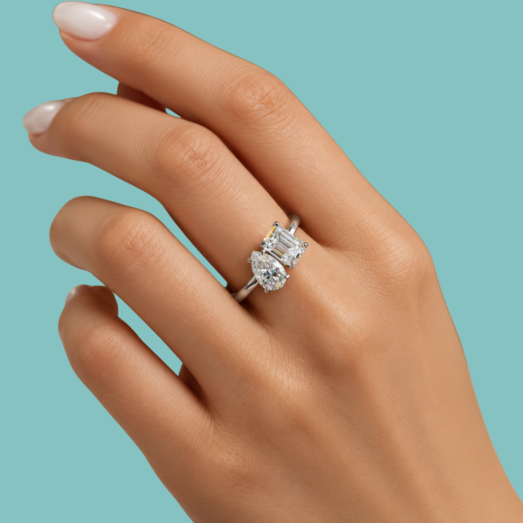 A close-up of a hand wearing a silver ring with a marquise-cut diamond and an emerald-cut diamond, set against a solid teal background.