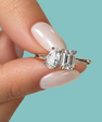 A close-up of a hand with pale pink manicured nails holding a silver engagement ring featuring a pear-cut diamond and an emerald-cut diamond, set against a solid light blue background.