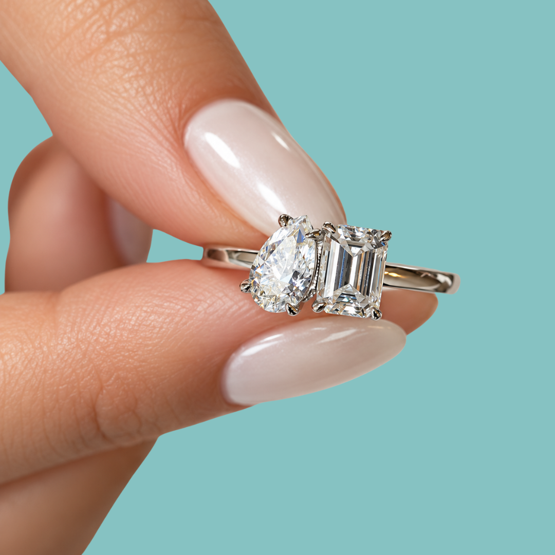 A close-up of a hand with pale pink manicured nails holding a silver engagement ring featuring a pear-cut diamond and an emerald-cut diamond, set against a solid light blue background.