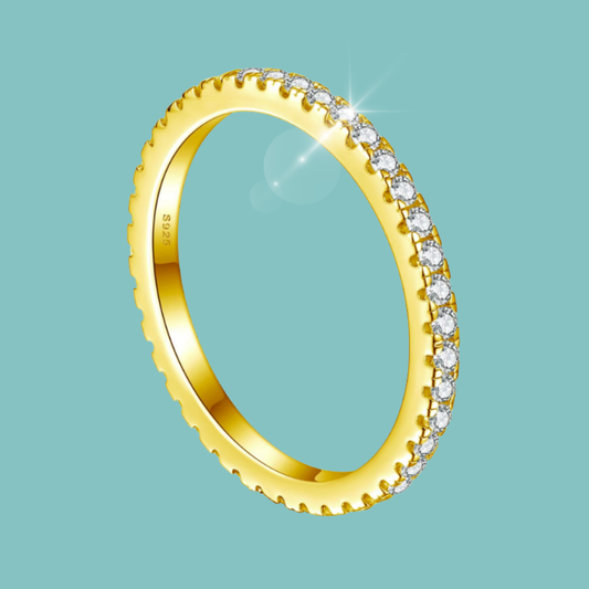 A yellow gold eternity ring featuring a row of clear round-cut gemstones on one side and a scalloped edge on the other, shown on a solid light teal background.