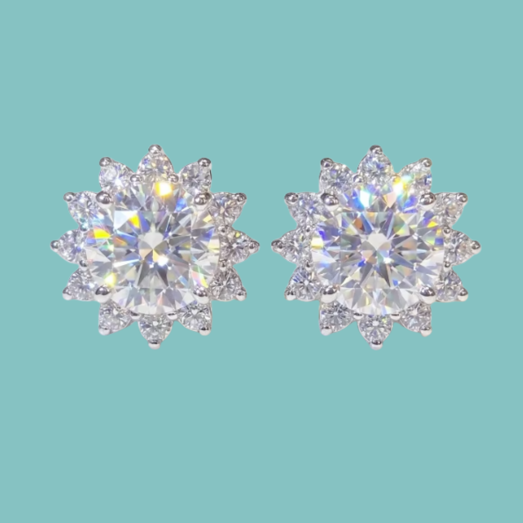 A pair of round-cut stud earrings with surrounding starburst clusters of small clear stones, shown on a solid light aqua background.