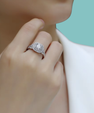A close-up lifestyle shot of a model wearing a silver-toned halo ring with a round-cut center stone and pavé band, set against a soft white shirt and a solid mint green background.