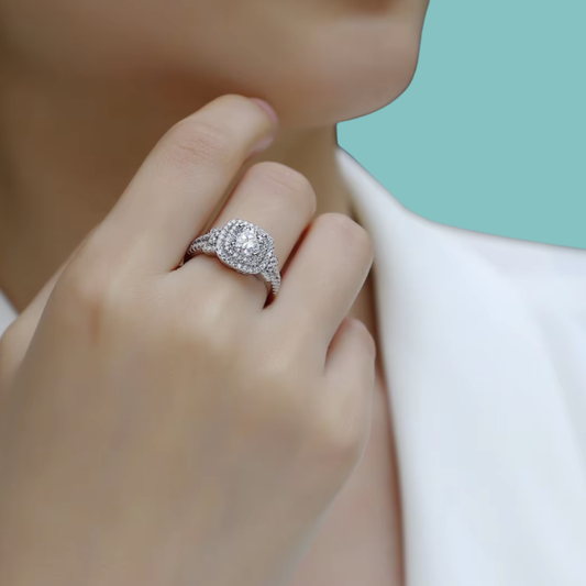 A close-up lifestyle shot of a model wearing a silver-toned halo ring with a round-cut center stone and pavé band, set against a soft white shirt and a solid mint green background.
