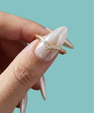 A close-up of a hand with long, glossy nude manicured nails holding a thin wavy gold ring adorned with small clear stones against a solid light teal background.