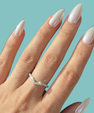 A close-up of a hand with long, glossy white manicured nails wearing a delicate silver V-shaped ring set with small sparkling stones, against a solid light teal background.