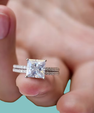 A close-up of a hand wearing a silver-toned ring set with a large princess-cut clear center stone and pavé-set side stones, against a soft teal background.