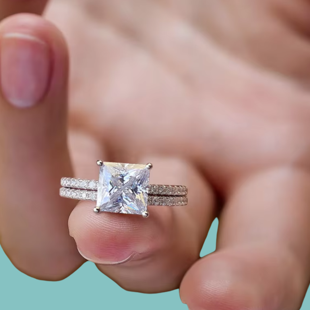 A close-up of a hand wearing a silver-toned ring set with a large princess-cut clear center stone and pavé-set side stones, against a soft teal background.