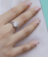 A close-up of a hand wearing a silver-toned ring with a heart-shaped clear gemstone and a matching band, resting on a white surface with a mint green corner visible.