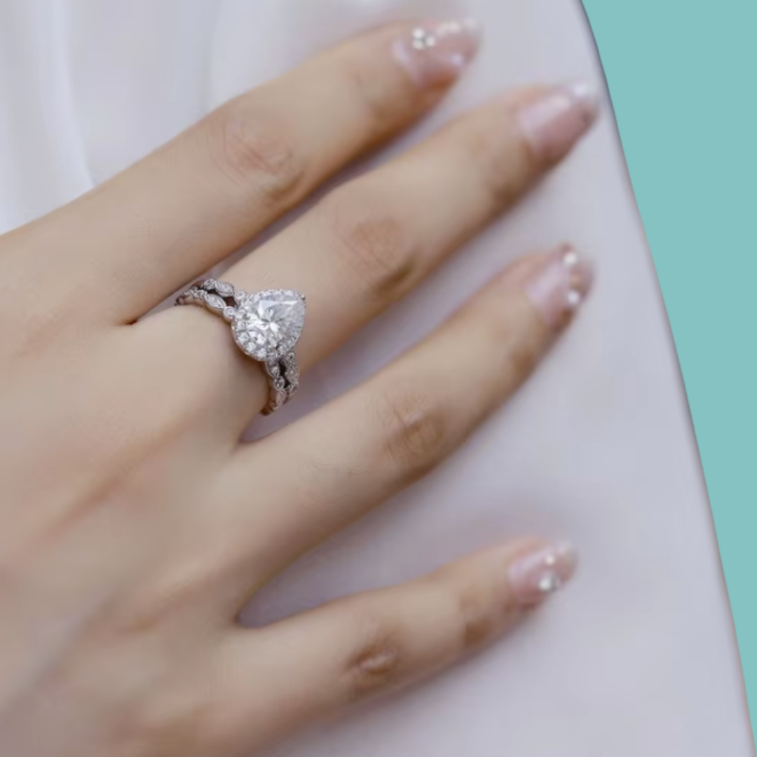 A close-up of a hand wearing a silver-toned ring with a heart-shaped clear gemstone and a matching band, resting on a white surface with a mint green corner visible.