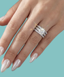 A close-up of a hand with long, glossy nude-pink manicured nails wearing a sparkling three-band diamond ring set against a solid teal background.