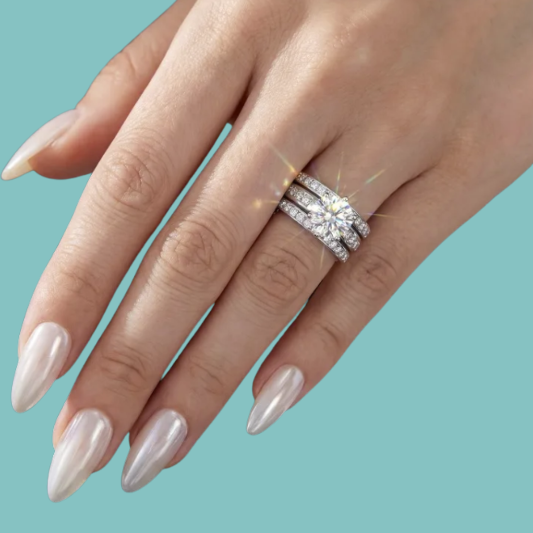 A close-up of a hand with long, glossy nude-pink manicured nails wearing a sparkling three-band diamond ring set against a solid teal background.