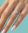 A close-up of a hand wearing a yellow gold ring with a round brilliant-cut diamond. The background is a solid light teal color.