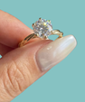 A close-up of a hand with long, glossy nude manicured nails holding a gold ring with a large round brilliant-cut diamond. The background is a solid light teal color.