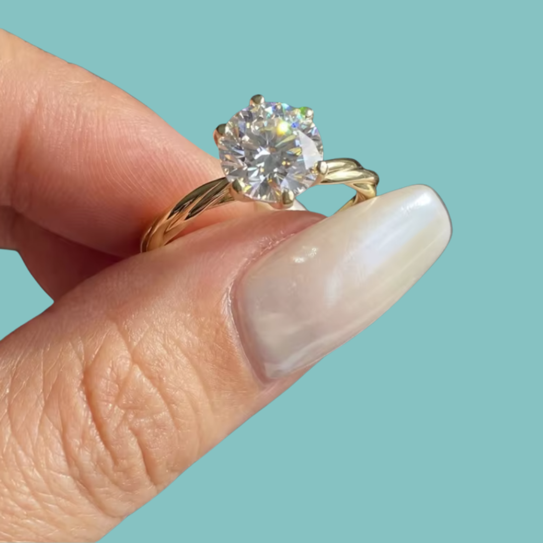 A close-up of a hand with long, glossy nude manicured nails holding a gold ring with a large round brilliant-cut diamond. The background is a solid light teal color.