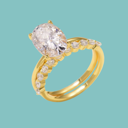 A yellow gold ring featuring a large oval-cut clear center stone and small clear accent stones along a split shank, shown on a solid light aqua background.