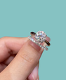 A close-up of a hand holding a silver ring set with a round brilliant-cut diamond and a matching diamond pavé band, against a solid light teal background.