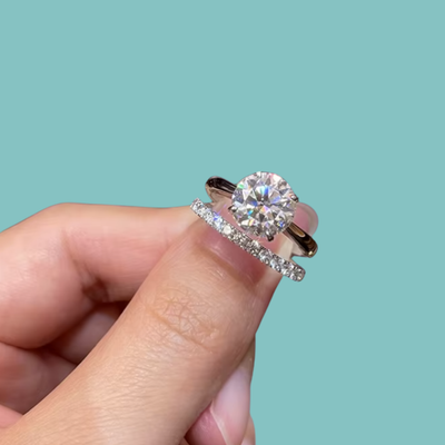 A close-up of a hand holding a silver ring set with a round brilliant-cut diamond and a matching diamond pavé band, against a solid light teal background.