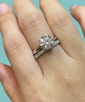 A close-up of a hand wearing a silver-toned engagement ring with a round brilliant-cut center stone and a matching pavé band, set against a solid light blue background.