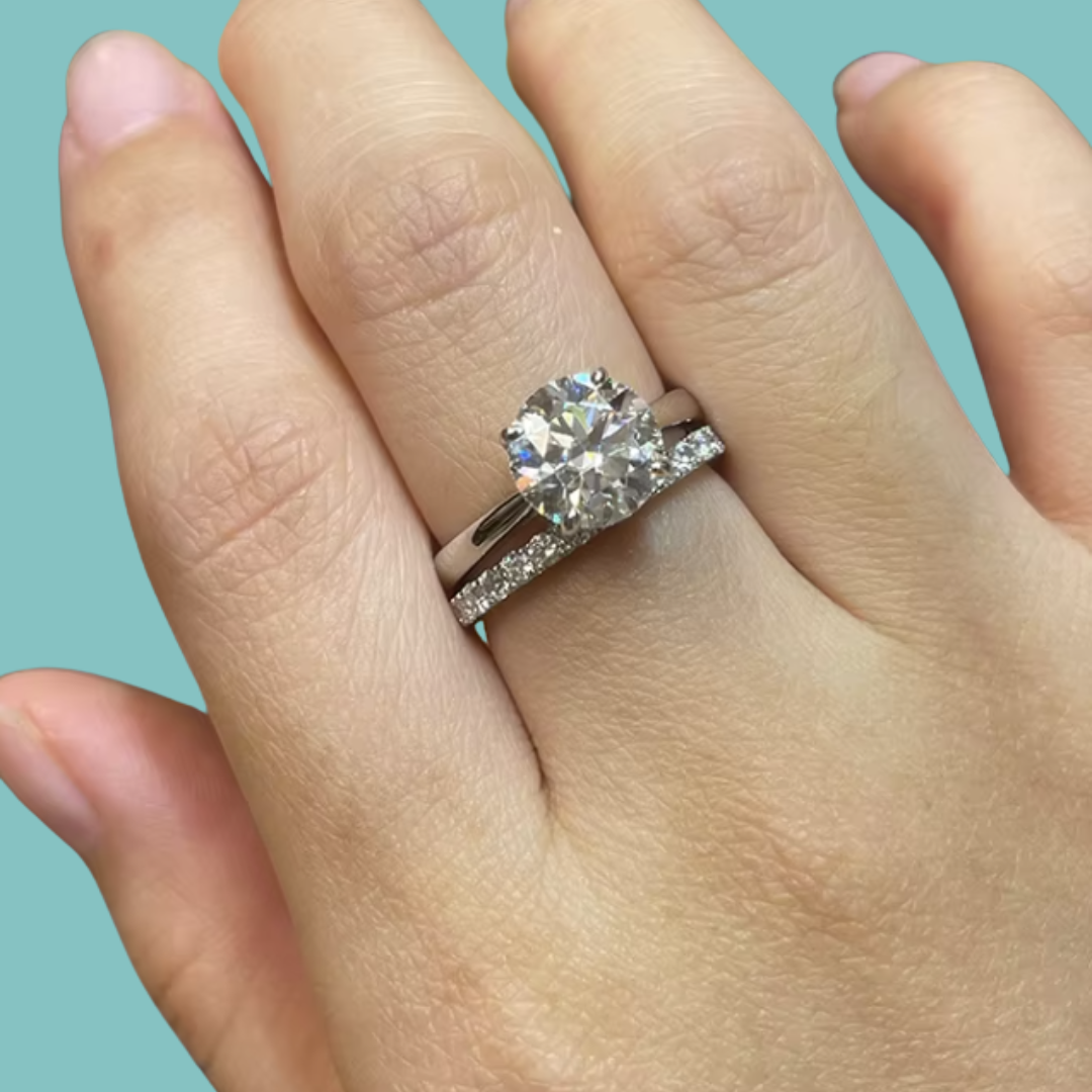 A close-up of a hand wearing a silver-toned engagement ring with a round brilliant-cut center stone and a matching pavé band, set against a solid light blue background.