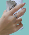 A silver ring with an oval moissanite gemstone is worn on a person's finger, with a clear faceted glass held behind the hand against a solid light aqua background.