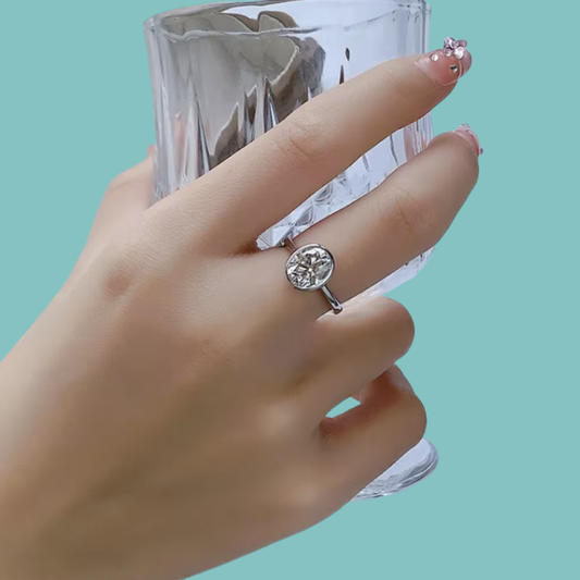 A silver ring with an oval moissanite gemstone is worn on a person's finger, with a clear faceted glass held behind the hand against a solid light aqua background.