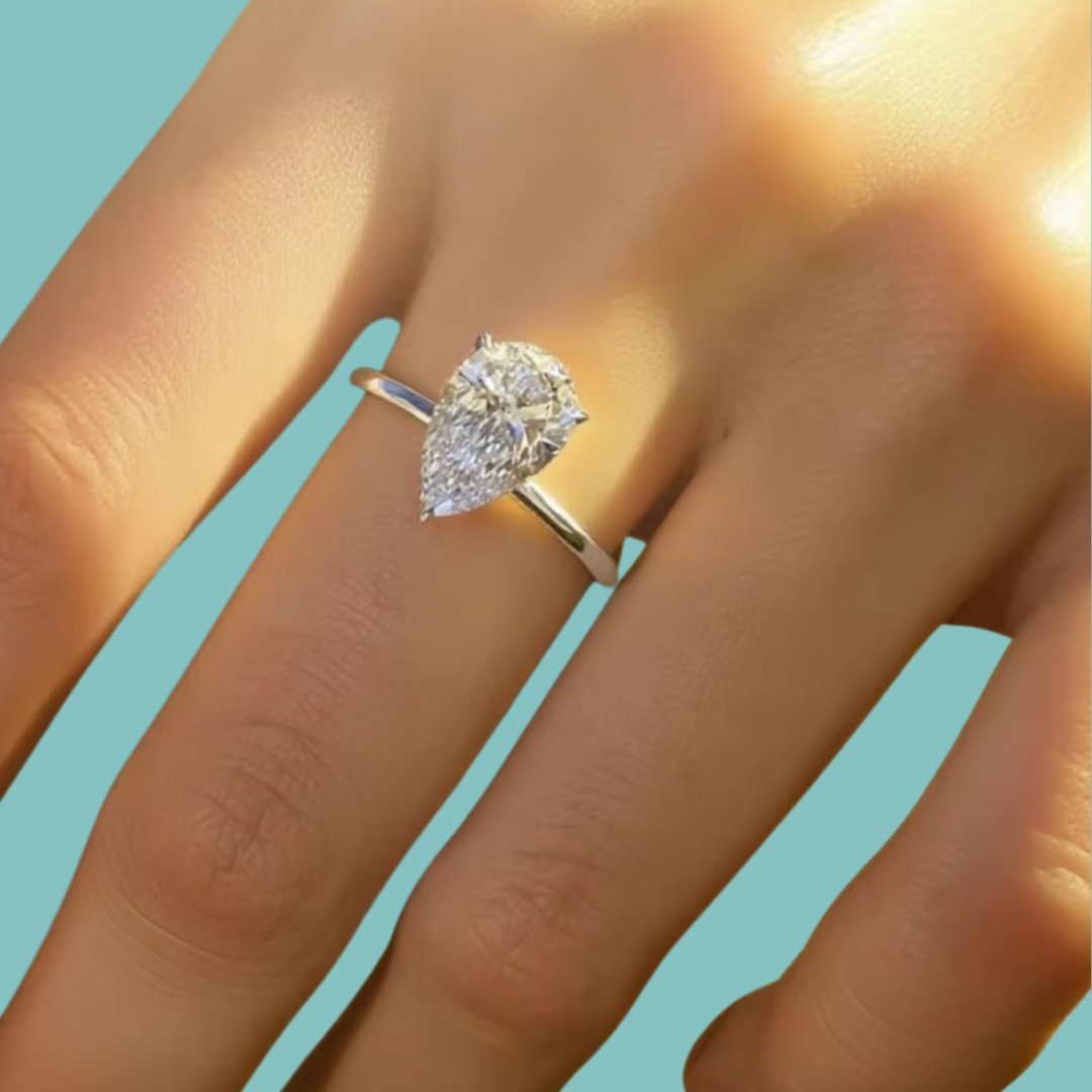 A pear-cut diamond ring on a silver band, shown worn on a person's finger against a solid light blue background.