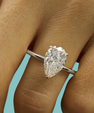 A pear-cut diamond ring on a silver band, shown worn on a person's finger against a light blue background.