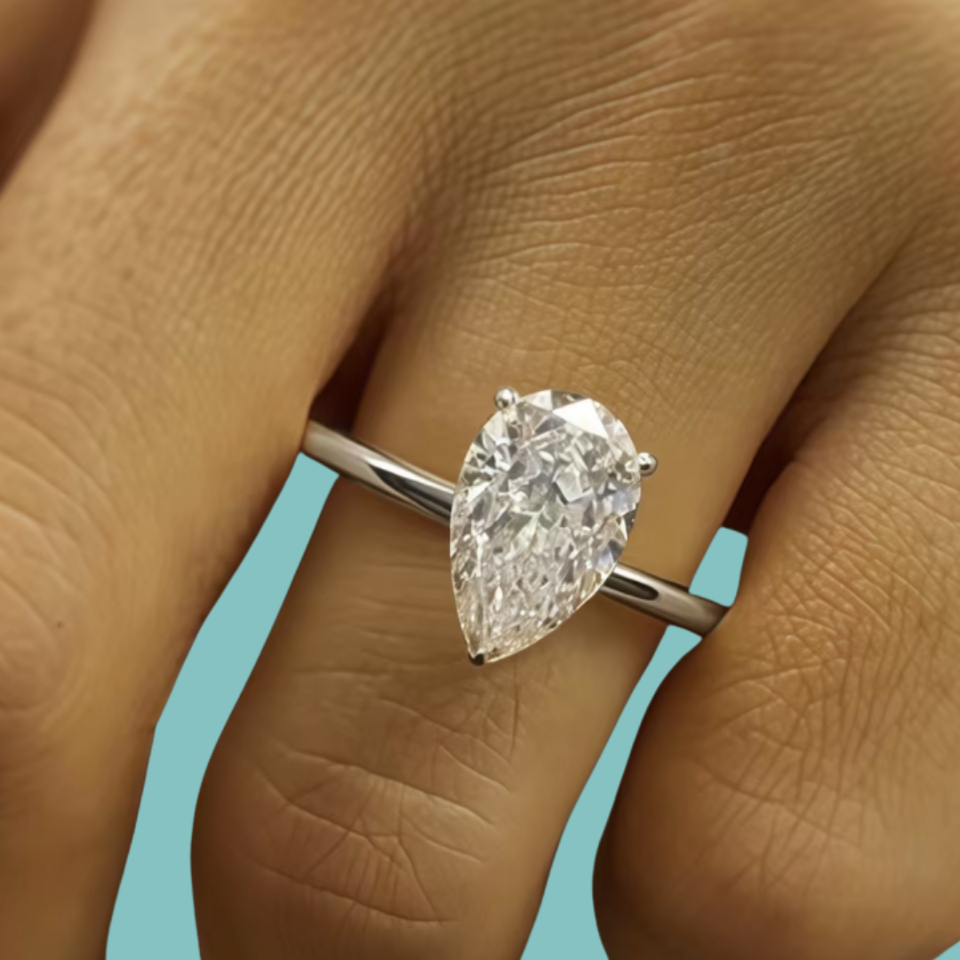 A pear-cut diamond ring on a silver band, shown worn on a person's finger against a light blue background.