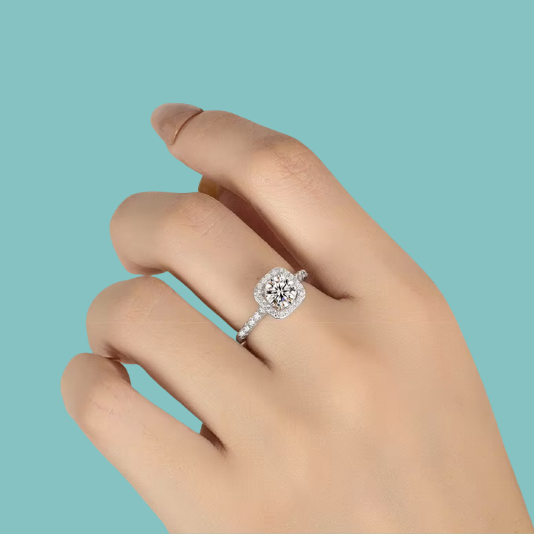 A silver-toned halo ring with a round-cut center stone and small accent stones, shown worn on a hand against a solid light blue background.