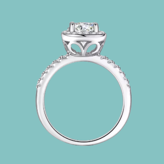 A silver-toned diamond ring with a round-cut center stone and a halo setting, shown against a solid light teal background.