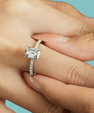 A silver engagement ring with an emerald-cut clear center stone and a pavé band, shown worn on a person's finger against a solid light blue background.