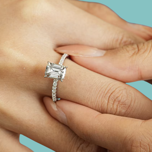 A silver engagement ring with an emerald-cut clear center stone and a pavé band, shown worn on a person's finger against a solid light blue background.