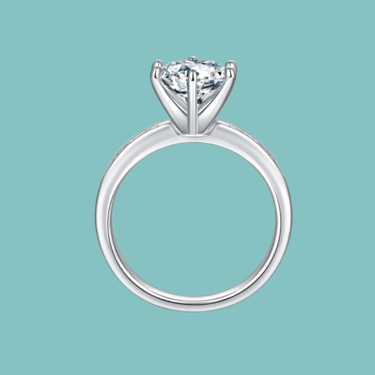 A silver solitaire engagement ring with a round brilliant-cut diamond, shown against a solid light teal background.