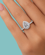 A pear-shaped halo ring with a pavé band, shown worn on a hand against a solid light blue background.