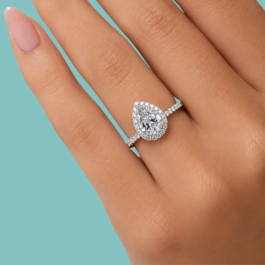 A pear-shaped halo ring with a pavé band, shown worn on a hand against a solid light blue background.
