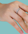 A close-up of a hand wearing a thin silver band ring set with small clear stones, against a solid light aqua background.