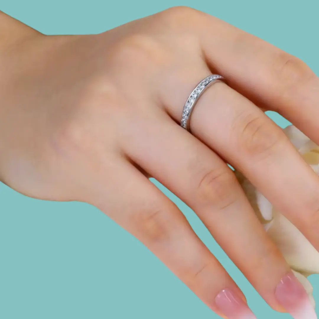A close-up of a hand wearing a thin silver band ring set with small clear stones, against a solid light aqua background.