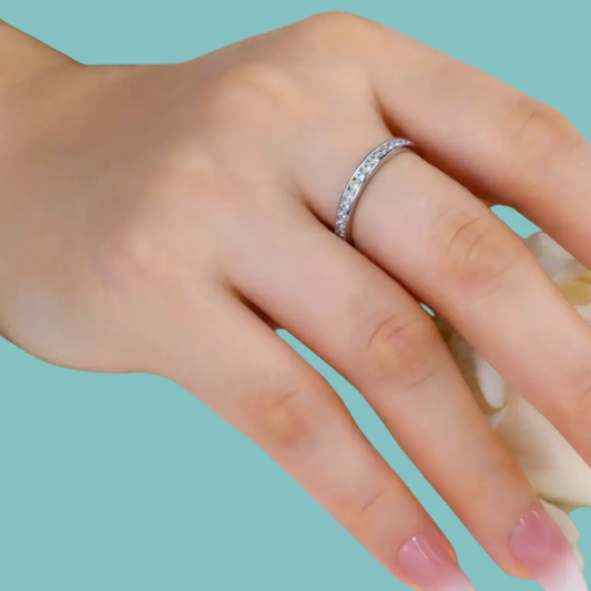 A close-up of a hand wearing a thin silver band ring set with small clear stones, against a solid light aqua background.